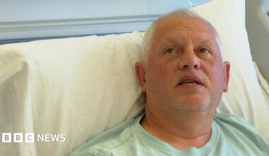 A man with grey hair wearing a light green t-shirt lies in a hospital bed, propped up against a pillow. He is looking upwards.