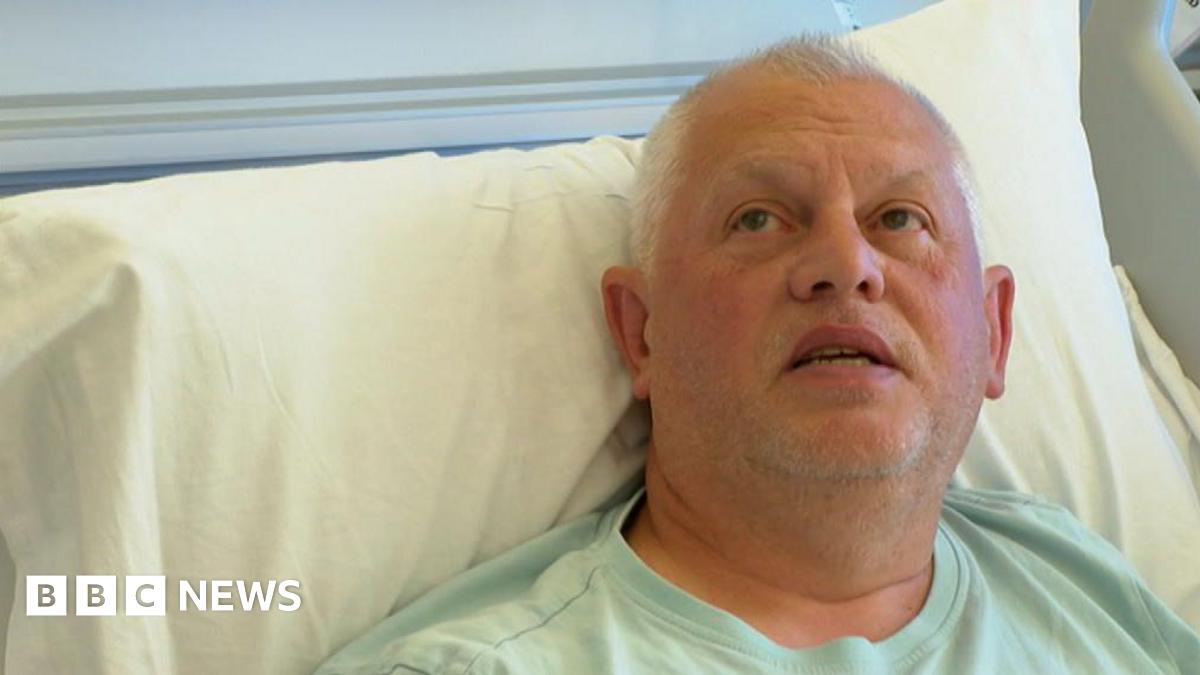 A man with grey hair wearing a light green t-shirt lies in a hospital bed, propped up against a pillow. He is looking upwards.