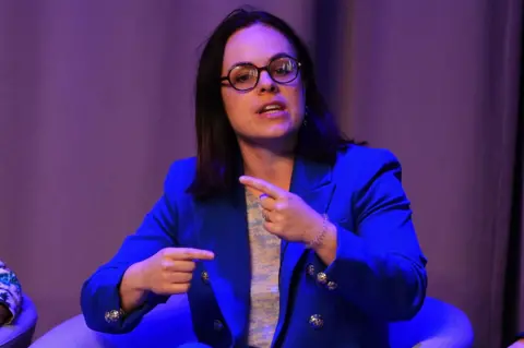 PA Media Kate Forbes speaks on stage. She sits in an armchair and wears a blue blazer, black glasses and gestures with her hands.