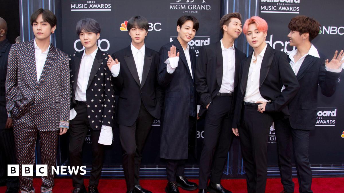 The seven members of BTS line up on a red carpet at the 2019 Billboard Music Awards. Each wears a tailored suit - two members wear patterned jackets. One member has his hair dyed salmon pink.