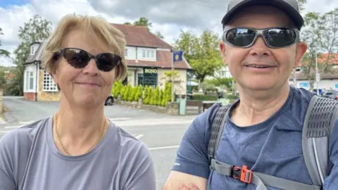 Gemma Dillon/BBC A man and woman are standing with a busy main road in the background. 
The woman has short blonde hair and is wearing sunglasses.
The man has a grey hat and sunglasses. He is wearing a blue t-shirt and carrying a rucksack. 