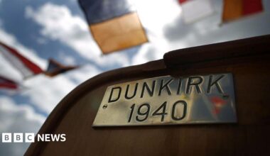 A brass plate reading "DUNKIRK 1940" on the deck of a boat. In the background there are semaphore flags on bunting.