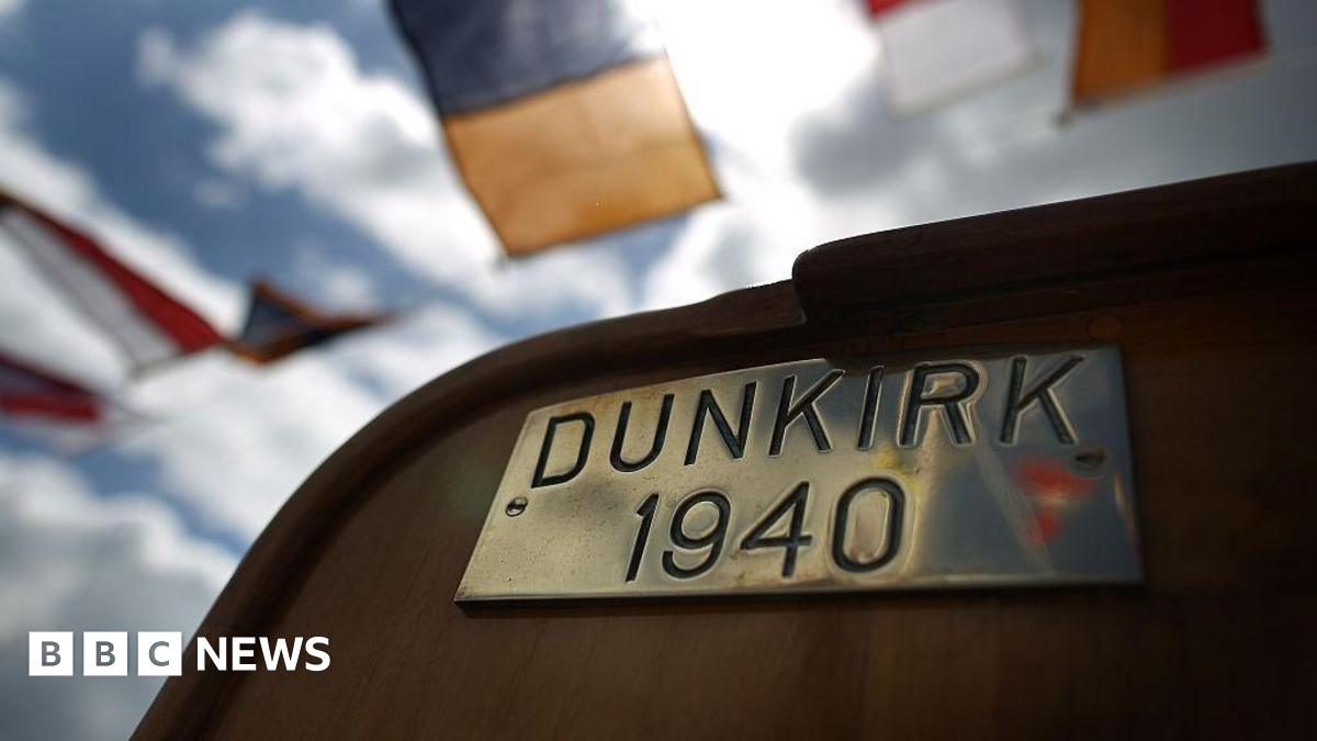 A brass plate reading "DUNKIRK 1940" on the deck of a boat. In the background there are semaphore flags on bunting.