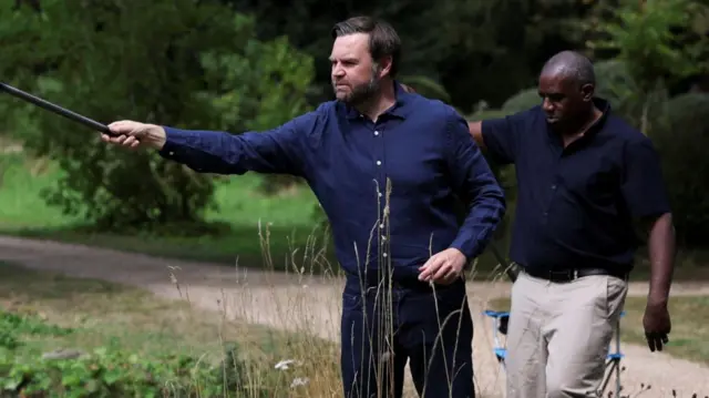 JD Vance stands in a dark blue shirt and black trousers as he holds up a fishing rod on the banks of a small lake, David Lammy in a short-sleeved dark shirt and tan trousers standing behind them