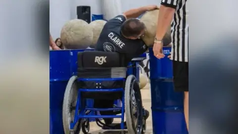 Gary Clarke Mr Clarke in his wheelchair and his back to the camera as he lifts weighted balls. He is wearing a navy blue t shirt with his surname Clarke written on it in white writing. There is what looks like an umpire beside him wearing a black and white striped t shirt and black shorts. 