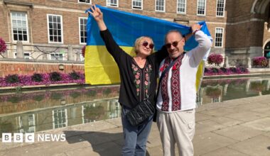 A woman and a man arm in arm, holding up a blue and yellow Ukrainian flag behind them as they stand on Bristol's College Green. They are both wearing Ukrainian-style embroidered clothes.