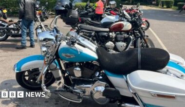 Various motorbikes queued up at a carpark in Micklesham, the font bike is blue and white with a dark coloured seat.