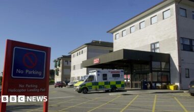 An ambulance is parked outside the entrance of a hospital building. A red no parking sign is on display, it says no parking, helicopter landing pad. The hospital building is grey.