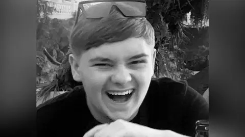 Facebook A black and white picture of the teenager who is laughing. He wears sunglasses on the top of his head, trees and plants can be seen in the background.