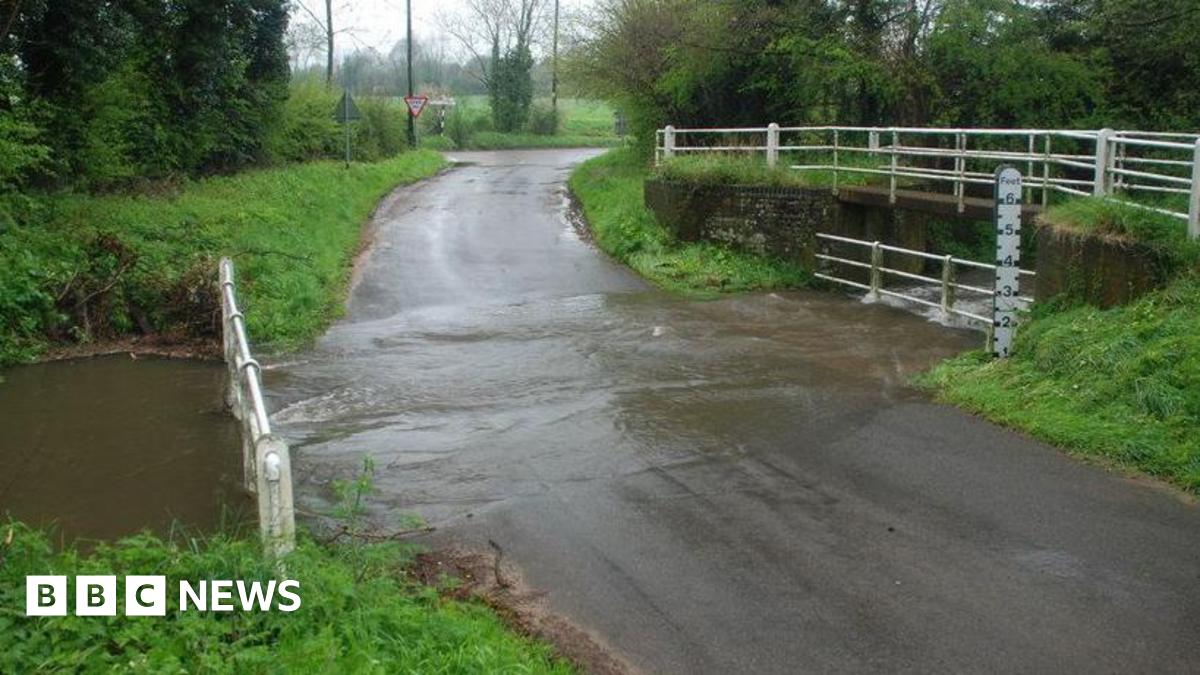 A ford with a river running over the road. Next to the ford is a bridge. There are white railings on both of them, trees on both sides of the road, and a road junction in the distance.