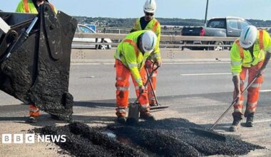 Three highway maintenance workers wearing hi-vis orange trousers, yellow hi-vis jackets and white hard hats. They are using tools to flatten a steaming pile of fresh tarmac, which is being poured into a fault in the road by a digger.