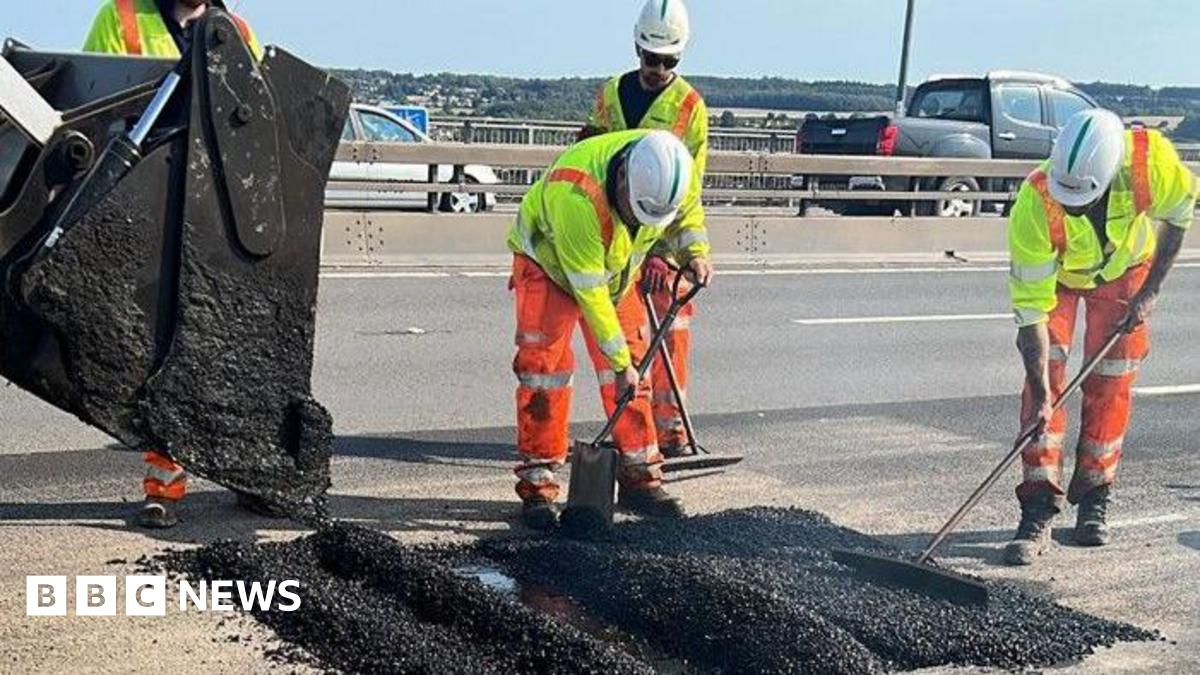 Three highway maintenance workers wearing hi-vis orange trousers, yellow hi-vis jackets and white hard hats. They are using tools to flatten a steaming pile of fresh tarmac, which is being poured into a fault in the road by a digger.