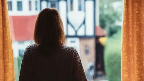 Getty Images Generic shot of the back of a woman with brown hair cur in a bib, wearing a dark-coloured woolen cardigan, looking out of a window towards a house with white render across much of the front with brown edging. The window of the woman's house has yellow curtains. A hedge is visible to the right.