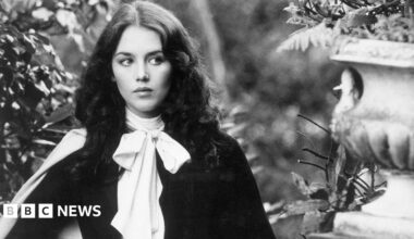 A black and white image of Isabelle Adjani who has brunette hair and is standing in a garden with stone ornaments.