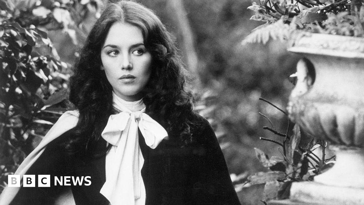 A black and white image of Isabelle Adjani who has brunette hair and is standing in a garden with stone ornaments.