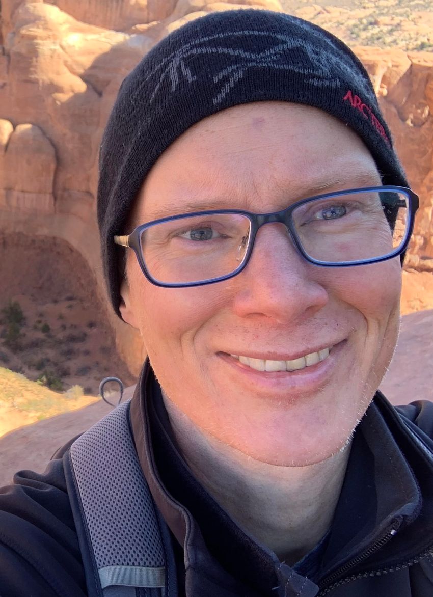 Close-up selfie image of Christopher Gregg, wearing glasses, dark patterned hat, a jacket, and a backpack in front of a geological formation.
