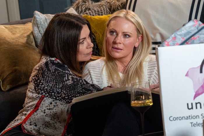 Carla and Lisa sitting on the floor with a glass of alcohol on the table above them in Coronation Street