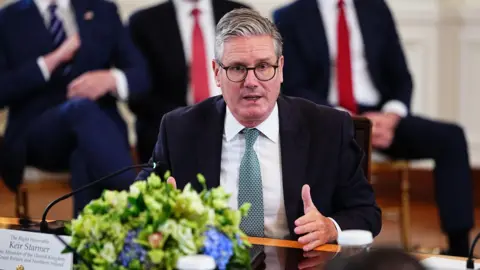 EPA Keir Starmer sitting at glossed wooden table in White House meeting with other leaders. The camera is on him as he speaks. Starmer is gesturing with his left hand and is wearing a black blazer, a white shirt and a patterned blue tie. There are blue flowers in front of him on the table. Behind, sit three men against the wall - the heads are out of shot, but they are wearing suits, white shirts and a stripy navy and white tie, a red tie and a red tie respectively from left to right. 