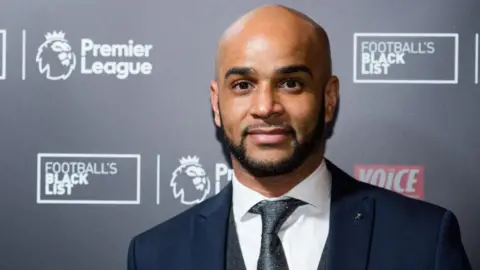 Getty Images Leon McKenzie is dressed in a white shirt, grey tie, grey waistcoat and dark blue jacket. He has a shaved head and a trimmed beard and moustache. He is standing in front of Premier League branded hoarding, with other imagery including a statement that reads: "Football's Black List".