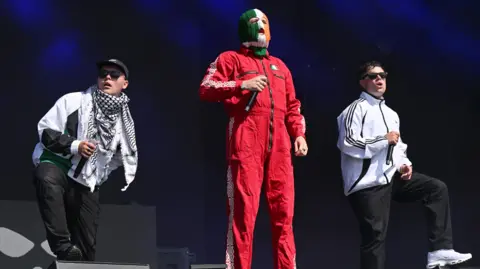 Leon Neal/Getty Images Mo Chara, DJ Próvaí and Móglaí Bap on stage at Glastonbury in June.  Mo Chara and Móglaí Bap are dressed in white sports jackets and black trousers and are each resting a foot on a speaker.  DJ Próvaí stands between them dressed in a red jumpsuit and a striped green, white and orange balaclava. 