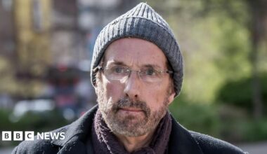 Chris Brain looks at the camera. He has a grey beard and is wearing glasses, a grey beanie hat and a dark coat and scarf.
