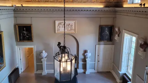 Supplied A view from above of a room with busts of Roman gods. There are ornate paintings hanging on the walls and a large lamp hanging from the ceiling. There are windows on the right hand side.