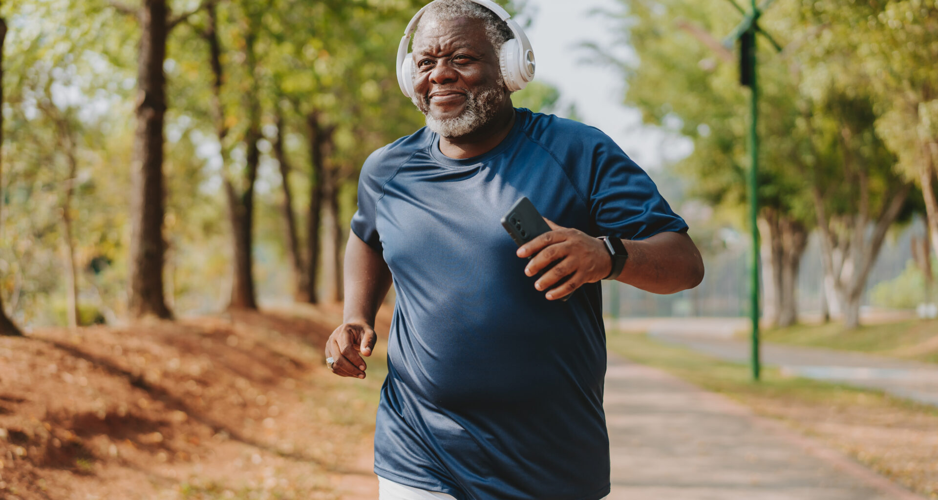 A man runs in a park, wearing a sports t-shirt and a fitness tracker. He has overear headphones on and holds his phone in his hand. Behind him we see a long row of leafy trees.