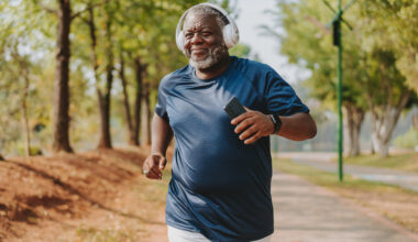 A man runs in a park, wearing a sports t-shirt and a fitness tracker. He has overear headphones on and holds his phone in his hand. Behind him we see a long row of leafy trees.