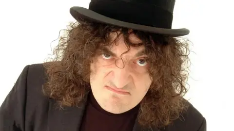 Jerry Sadowitz A man in a black blazer and top hat with long, brown curly hair - he raises one eyebrow and wears a grumpy expression
