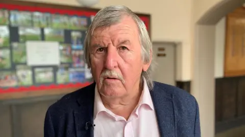 Iestyn Thomas, who has grey hair, is looking at the camera. He has a blue jacket and a light pink shirt. He also has a grey moustache.