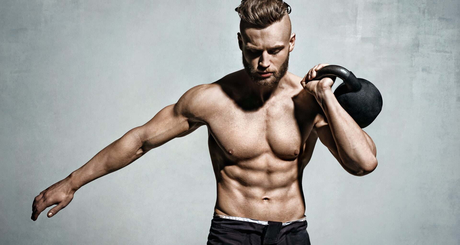 Man holding one of the best kettlebells up to his shoulder using left arm against a grey backdrop