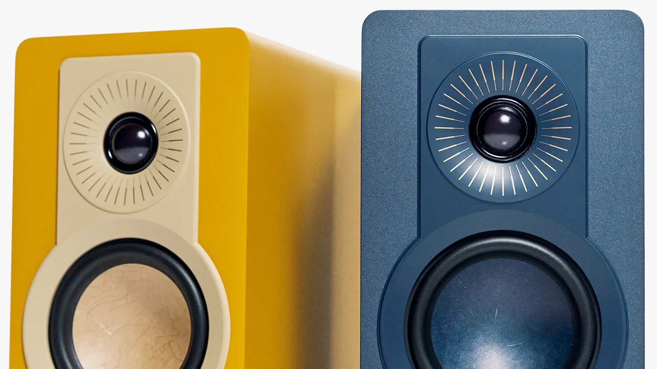 Two modern bookshelf speakers, one yellow with a beige front panel and one dark blue, each featuring a circular tweeter with radial lines and a larger woofer below.