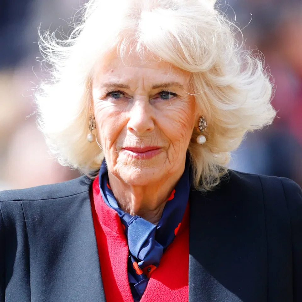  Queen Camilla wears a black blazer and a red dress and has blonde hair which is in an upheaval in the wind. 