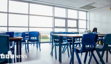 A generic image of a classroom - there are several desks with classroom chairs and a window in the background with a view of the countryside in the background.