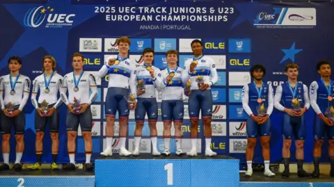 UEC Ioan with his team-mates in their full kit, standing in first place on the Track Juniors European Championships podium with their medals round their necks. Their cycling kit includes a white long-sleeved shirt and blue shorts. The second and third place teams are on their stands.  