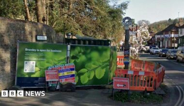 A google maps image showing green BT Openreach hoardings next to orange safety barriers at an engineering site in the centre of Bramley. Cars and a village sign can be seen in the background.