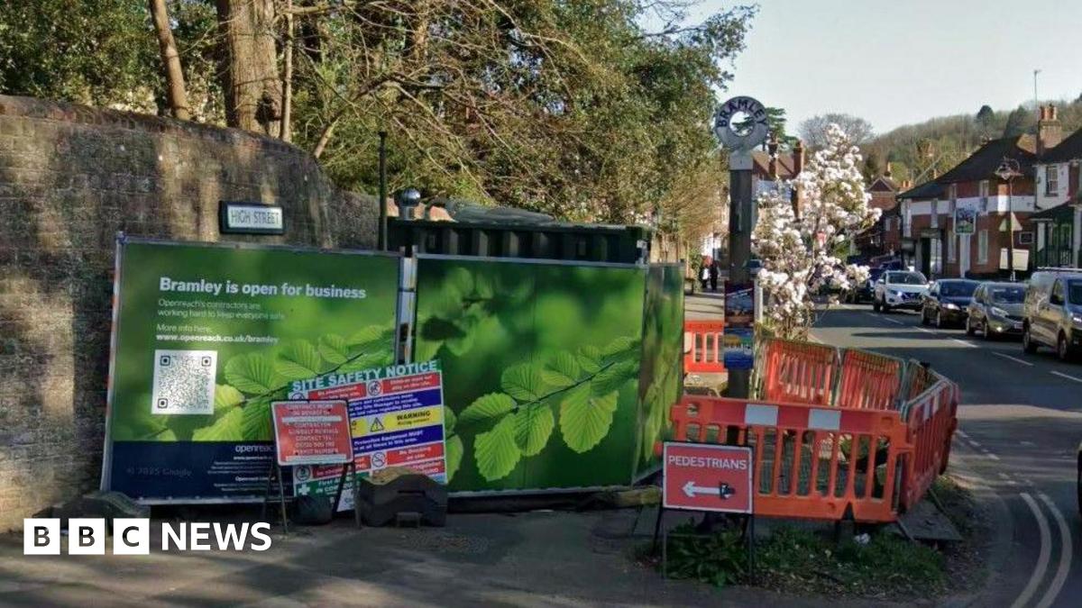 A google maps image showing green BT Openreach hoardings next to orange safety barriers at an engineering site in the centre of Bramley. Cars and a village sign can be seen in the background.