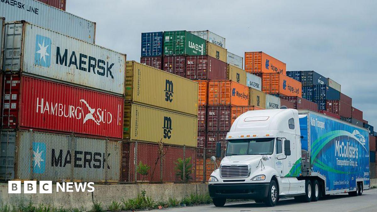 A blue and white McCollister's Transportation Group tractor trailer drives past a pile of shipping containers at the Port of Baltimore in Baltimore, Maryland, US, on Friday, Aug 1, 2025.