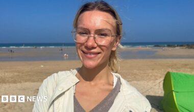 A woman with glasses wearing a light coloured coat and a grey sweater on a beach with a beach shelter behind her.