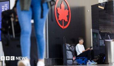 A middle-aged male passenger waits with his luggage at Toronto Pearson Airport. The legs of a woman in jeans can be seen blurred in the foreground. She is also pulling a suitcase.