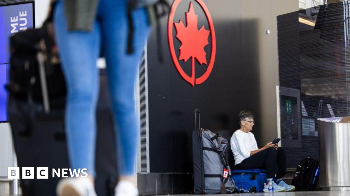 A middle-aged male passenger waits with his luggage at Toronto Pearson Airport. The legs of a woman in jeans can be seen blurred in the foreground. She is also pulling a suitcase.
