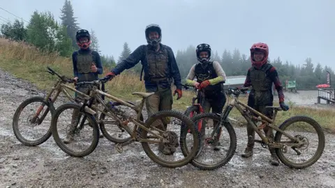 Family photo A group photo of the family at a wet trail centre, stood with their bikes. They are covered in mud head to toe. They're wearing full-face helmets but you can tell they're all smiling. 