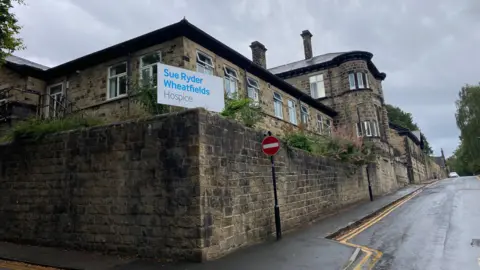 Cathy Minton/BBC A stone building on the corner of a road with a sign outside which reads 'Sue Ryder Wheatfields Hospice'.