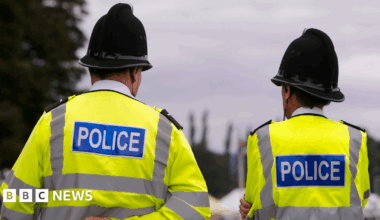 Two police officers stand with their backs to the camera, side by side in hi-vis jackets with 'POLICE' written on the back