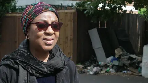 Animot Otunuga in a black outfit and a pink, orange and teal patterned hat. She stands in front of fly-tipped waste on the street she lives on in Thornton Heath. 
