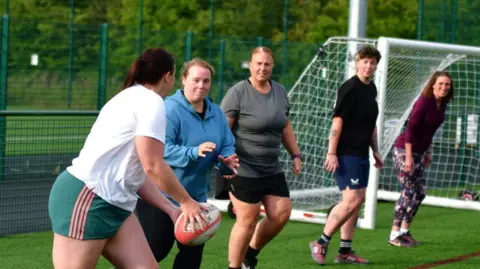 Washington Rugby Club A line of five women playing rugby. To the left, the player closest to the camera is about to pass the ball. The second player is waiting to catch it. The women are wearing shorts and t-shirts of various styles and colours.