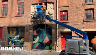 Two men on a cherry picker removing the bas-relief carving from the outside of the red brick Victorian building. Below them are two doorways covered in wooden panels painted with abstract artwork.