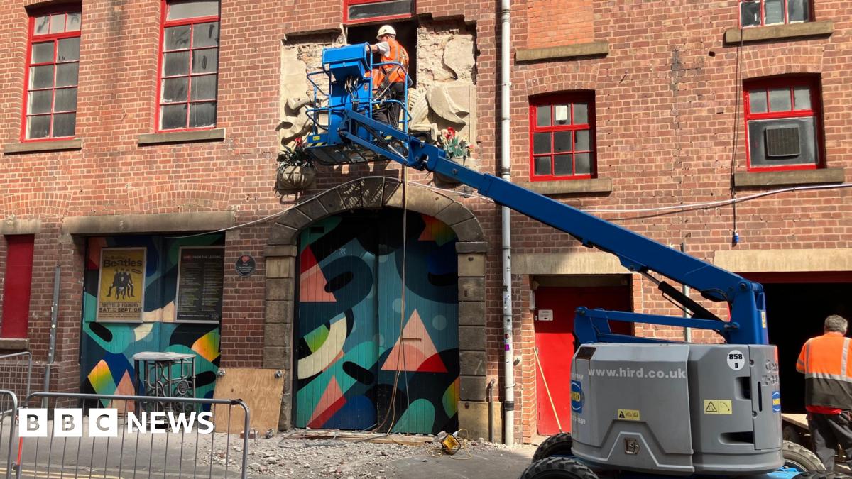 Two men on a cherry picker removing the bas-relief carving from the outside of the red brick Victorian building. Below them are two doorways covered in wooden panels painted with abstract artwork.