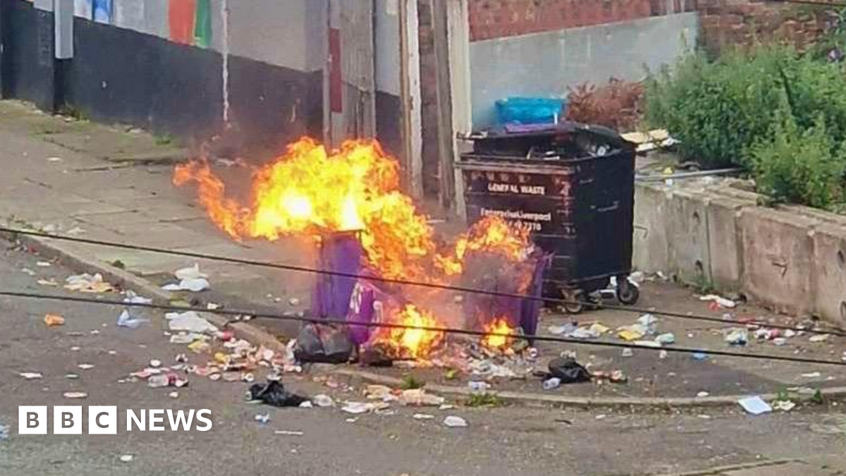 A wide shot of Sylvester Road in the Vauxhall area of Liverpool. There is a purple bin on fire surrounded by litter strewn across the pavement and road.