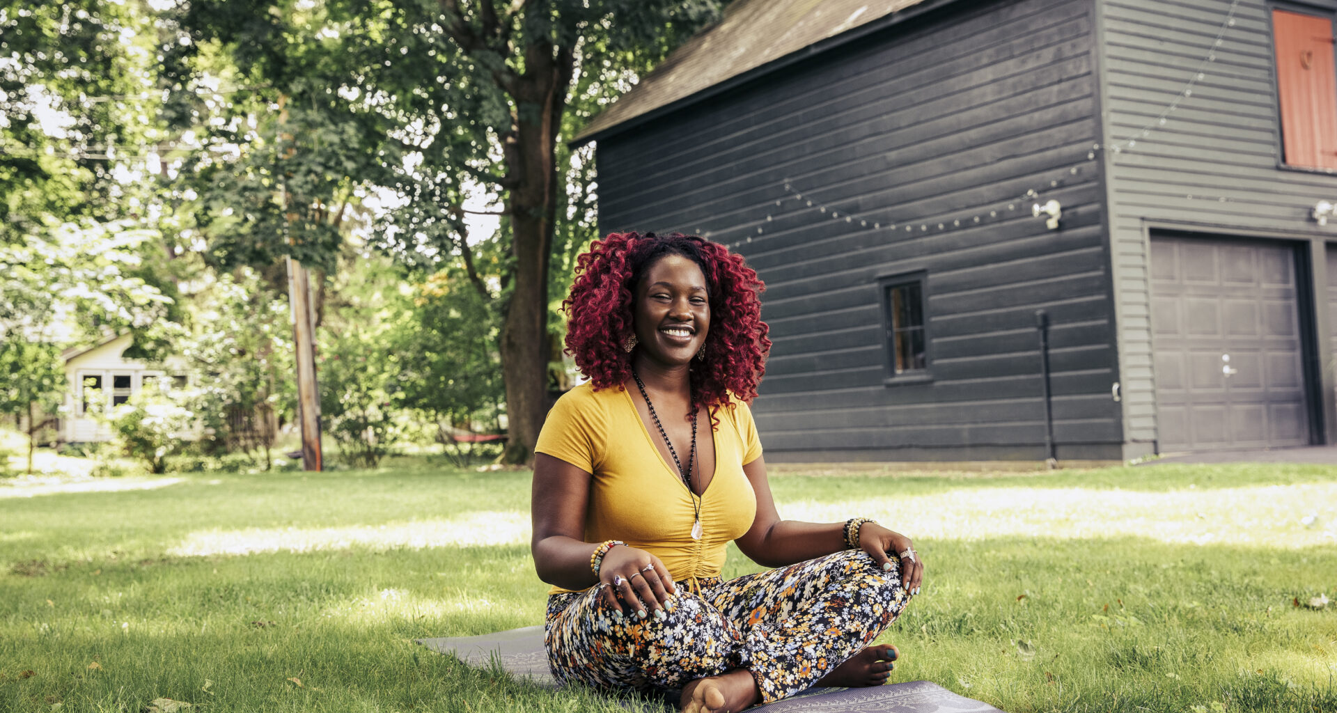 A woman sits cross-legged outside on a yoga mat. Behind her we see a large expanse of healthy-looking grass and the side of a house with a garage, which is decorated with fairy lights.